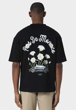 Mann iført svart t-skjorte med hvite blomster og grafikk av vintagebil, teksten "Pas De Monac" på ryggen, beige bukser, står med ryggen til.