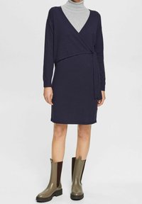 Marinblå omslagsklänning med långa ärmar och sjalkrage, tillsammans med gröna vattentäta ank stövlar med bruna detaljer och texturerade sidor.