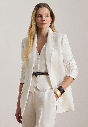 Kobieta ubrana w dopasowaną białą marynarkę, dopasowaną kamizelkę i spodnie, z plecionym paskiem oraz dużymi złotymi i żółwiowymi bransoletkami, pozująca z jedną ręką w kieszeni.