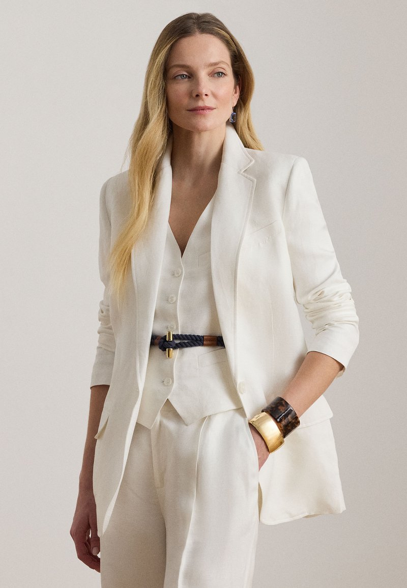 Γυναίκα που φοράει προσαρμοσμένο λευκό σακάκι, ταιριαστό γιλέκο και παντελόνι, μια πλεγμένη ζώνη και μεγάλα χρυσά και χελώνια βραχιόλια, ποζάροντας με το ένα χέρι στην τσέπη.