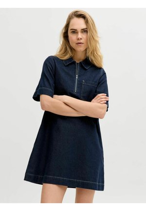 Femme aux longs cheveux blonds portant une robe en jean bleu foncé à manches courtes avec une fermeture éclair et une poche sur le devant, bras croisés, fond neutre.