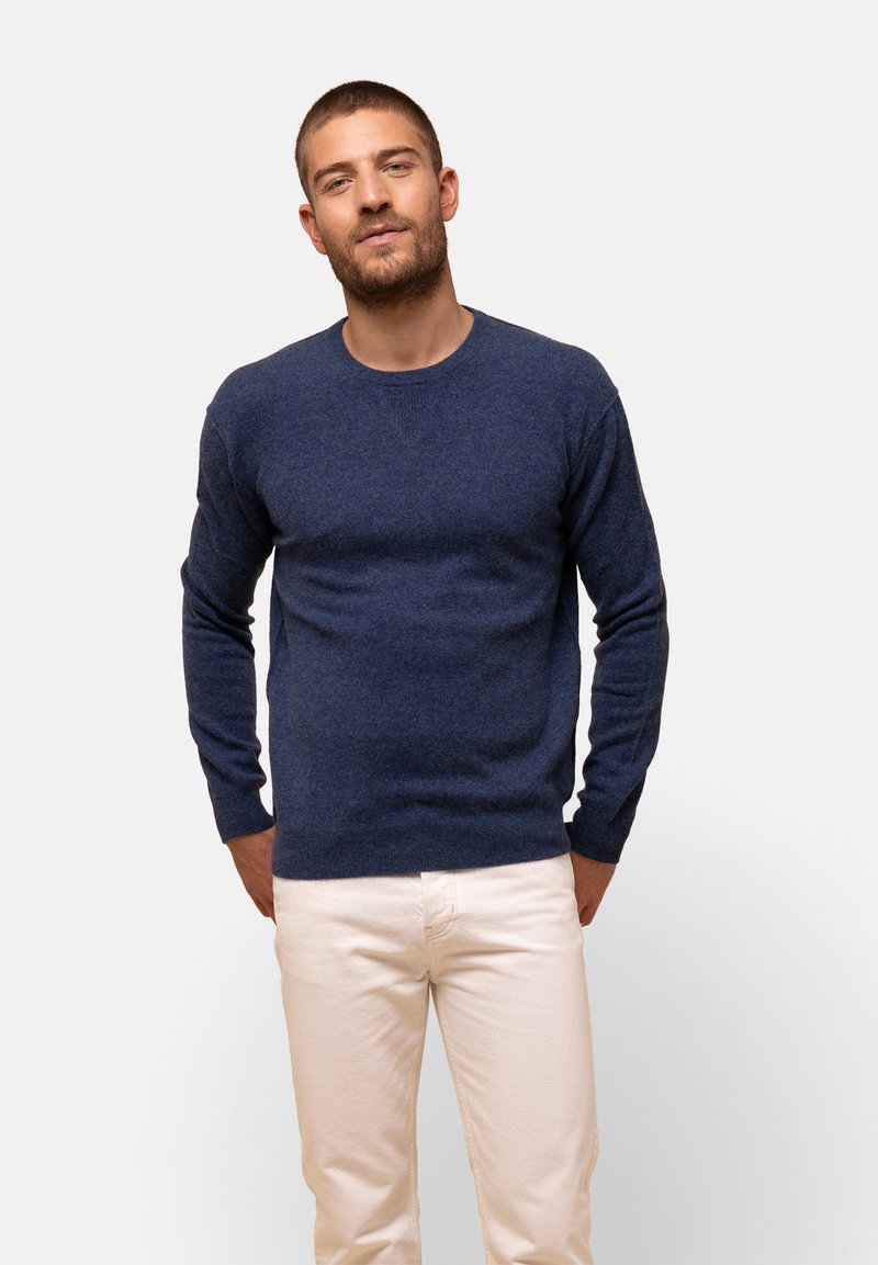 Ciemnoniebieski sweter z miękkiego dzianinowego materiału, z długimi rękawami i okrągłym dekoltem, w połączeniu z jasnymi spodniami. Prosty fason, bez wzorów.