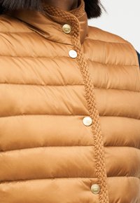 Καφέ φουσκωτό γιλέκο με πλεγμένο λαιμόκοψη, χρυσά κουμπιά και οριζόντιο σχέδιο καπιτονέ. Λείο υφή με ελαφρώς γυαλιστερό φινίρισμα.