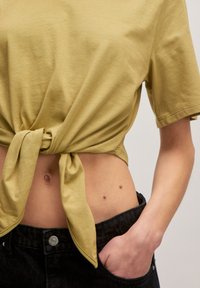 Licht olijfgroene katoenen crop top met een geknoopte taille; korte mouwen en een gladde textuur. Gecombineerd met zwarte spijkerbroek.
