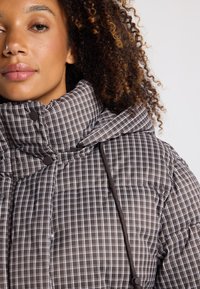 Chaqueta acolchada de cuadros marrón y blanco con cuello alto, que presenta drawstrings marrones y acentos de botones a presión. Textura suave y acolchada.