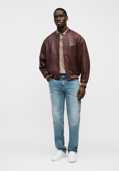 Καφέ δερμάτινο μπουφάν τύπου bomber με ριμπ μανσέτες, κεντημένο λογότυπο, συνδυασμένο με ανοιχτό μπλε distressed τζιν και λευκά αθλητικά παπούτσια.