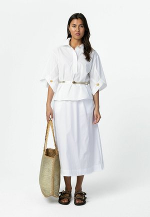 Žena v bielej blúzke s opaskom a sukni, drží tkanú tašku na rameno, nosí čierne sandále s ozdobnými cvočkami, stojí na jednoliatom bielom pozadí.