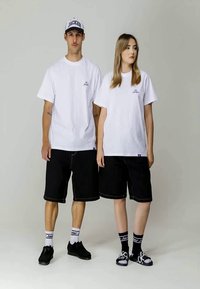 Camisetas de algodón blancas con pequeños logotipos bordados, pantalones cortos de mezclilla negros, zapatillas negras y calcetines en contraste; estilo casual y unisex.
