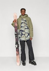 Pessoa com uma jaqueta de esqui verde e estampada segurando um esqui rosa e preto, vestindo calças de esqui pretas e botas de esqui pretas.