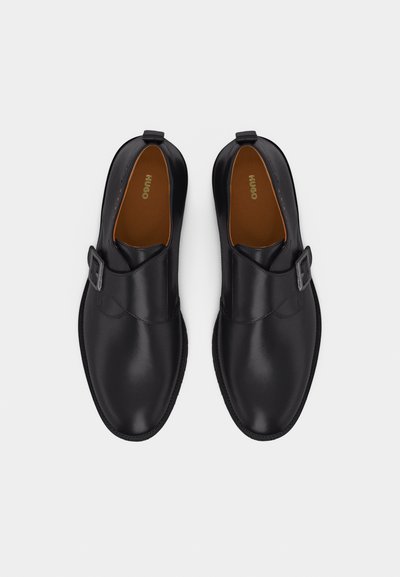 Paire de chaussures monastères noires en cuir avec boucles argentées et semelles intérieures beige, vues de dessus sur un fond blanc.