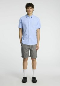 Camicia azzurra a maniche corte con colletto, abbinata a pantaloni corti grigi a scacchi, calzini bianchi e mocassini neri.