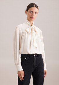 Blouse créme avec un col noué, de longues manches amples et des détails froncés au niveau de la poitrine ; associée à un jean noir ajusté.