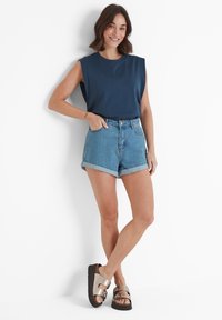 Woman wearing sleeveless navy top, rolled denim shorts, and metallic buckle sandals, standing with one hand in pocket against white wall.