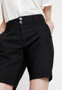 Czarne dopasowane szorty o gładkiej fakturze z dwoma guzikami, bocznymi kieszeniami i nowoczesnym krojem. Zestawione z białą koszulą na guziki.