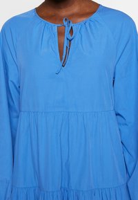Robe bleue à manches longues, ample, avec des volants froncés et un petit décolleté en V devant, noué avec des cordons bleus assortis.