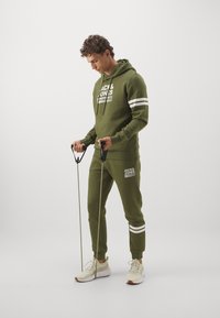 Olivgrüner Trainingsanzug mit weißen Akzentstreifen, bestehend aus einem Kapuzenpullover und schmal zulaufenden Hosen. Das Model hält Fitnessbänder und trägt helle Turnschuhe.
