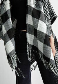 Poncho a scacchi bianco e nero con bordi frangiati, texture tessuta e un motivo pied-de-poule a contrasto su un lato.
