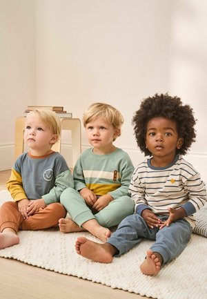 Tre barn sitter på en texturerad beige matta, klädda i färgglada bomullspyjamasar: blå, grön och randiga mönster med detaljer och grafiska tryck.
