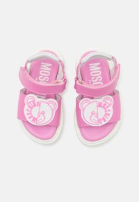 Růžové sandály s nastavitelnými popruhy, texturovaným povrchem a bílým logem medvěda na přední straně. Bílé podrážky s mírnou hloubkou.