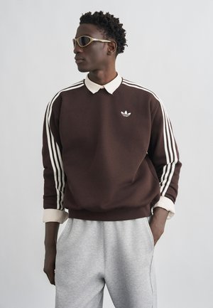 Mężczyzna ubrany w brązową bluzę Adidas z białymi paskami i logo, beżową koszulę z kołnierzykiem pod spodem, szare spodnie dresowe i okulary przeciwsłoneczne, patrzący w lewo.