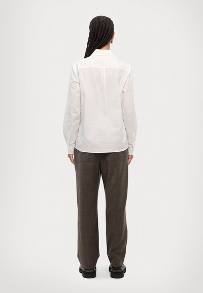 Long-sleeve white shirt with buttoned cuffs, back yoke detail. Paired with loose, brown check-patterned trousers and black platform shoes.