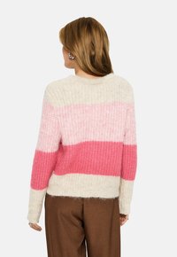 Kvinna i en texturerad stickad tröja med krämfärgade, ljusrosa och klarröda horisontella ränder, tillsammans med bruna byxor, vänd bort.