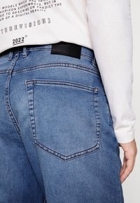 Jeans de ganga azul claro com um bolso traseiro com costura contrastante e uma etiqueta de couro preta na cintura. Usados com uma camisa de manga longa branca.