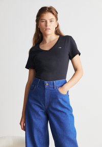 Femme aux longs cheveux portant un T-shirt noir à col en V et un jean bleu taille haute à jambes larges, une main dans la poche, fond neutre.