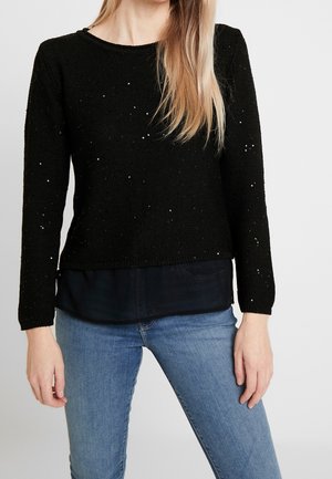 Kvinde iført sort langærmet paliettrøje over sort bluse kombineret med lyseblå jeans, stående mod en ensfarvet baggrund.