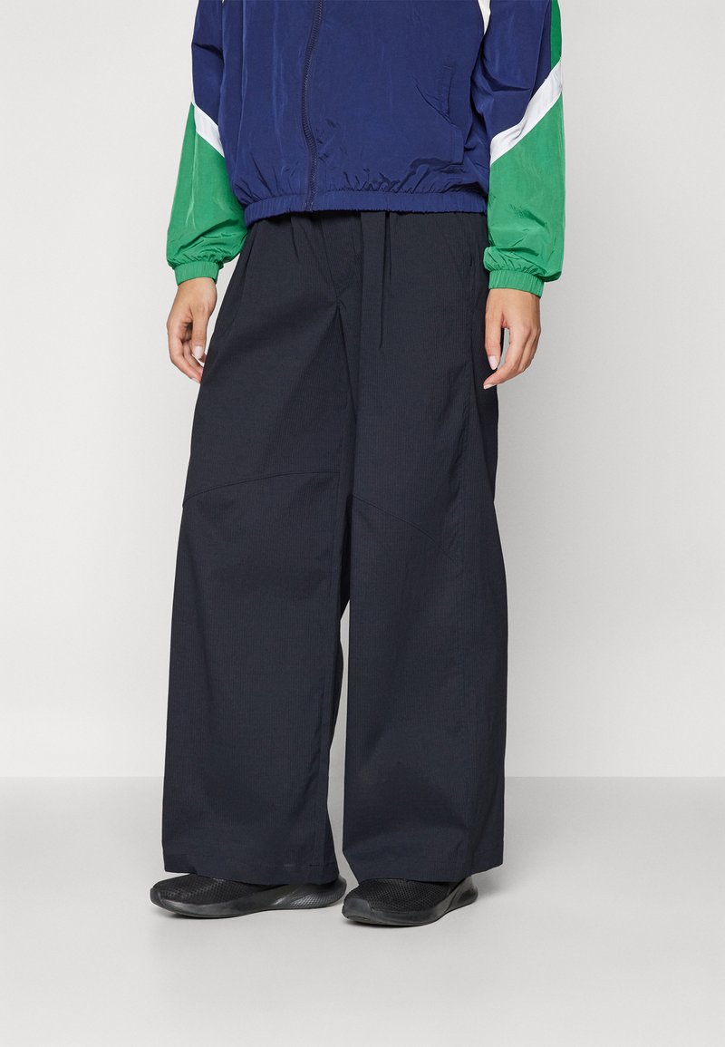 Pantaloni a gamba larga di colore blu navy in un tessuto testurizzato, con vita regolabile e tasche laterali, abbinati a un giubbotto blu e verde.