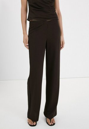 Žena v čiernych širokých nohaviciach a čiernom tielku bez rukávov, stojaca bosá na jednofarebnom bielom pozadí.