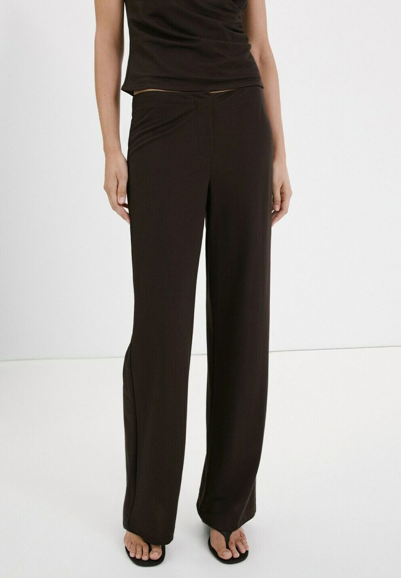 Femme portant un pantalon noir à jambes larges et un top noir sans manches, debout pieds nus sur un fond blanc uni.