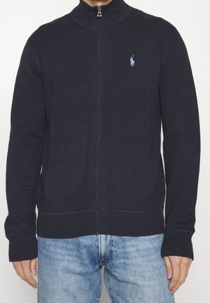 Homme portant un pull à fermeture éclair intégrale bleu marine avec un petit logo joueur de polo bleu clair sur la poitrine et un jean en denim bleu clair.