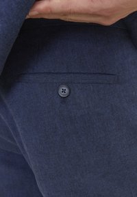 Detaliu al unei jachete de costum din in navy, având un singur buzunar pe spate cu un nasture închis la culoare și o textură subtilă. Materialul pare neted, cu o finisare mată.