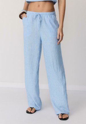 Svetlomodré voľné ľanové nohavice s pásom na sťahovanie, nosené s čiernymi sandálmi, odkryté bruško a jedna ruka v vrecku.