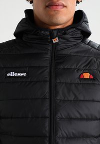 Schwarze Daunenjacke mit durchgehendem Reißverschluss, ausgestattet mit einer Kapuze und vertikaler Steppung. Enthält ein gesticktes Logo auf der Brust.