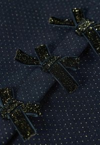 Tejido marino con pequeños puntos dorados, complementado por tres lazos de terciopelo oscuro y brillante. Textura suave con acentos decorativos.