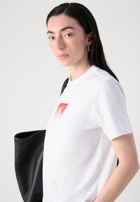 Witte katoenen t-shirt met een verlooplogo in rood en roze op de voorkant. Model houdt een zwarte leren schoudertas vast, met een effen achtergrond.