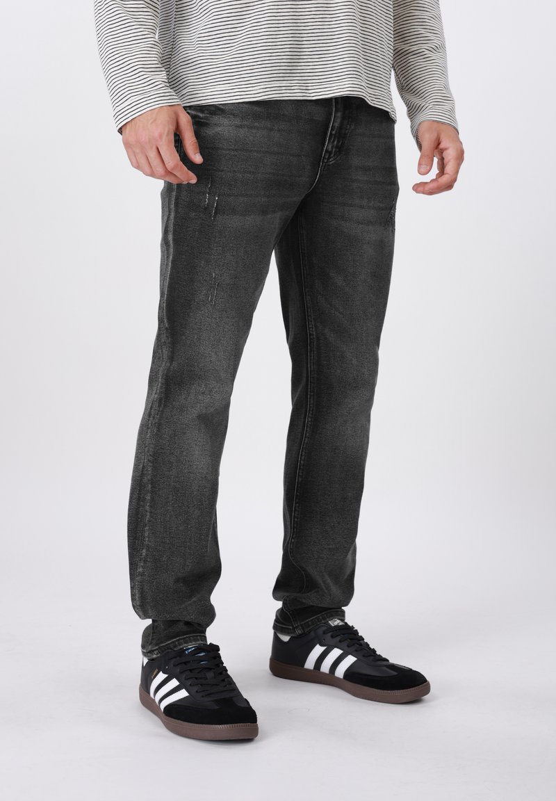 Μαύρα τζιν από τζιν με ελαφρύ ξεθώριασμα και φθαρμένες λεπτομέρειες, σε συνδυασμό με μαύρα αθλητικά παπούτσια με λευκές ρίγες και καφέ ελαστική σόλα.