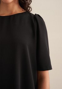 Schwarze Kurzarm-Bluse aus glattem Stoff, mit lockerer Passform und dezentem Puffdetail an den Schultern. Schlichter Ausschnitt, minimalistisches Design.
