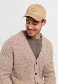Cappellino da baseball beige con logo ricamato, abbinato a un cardigan lavorato a maglia rosa chiaro con scollatura a V e bottoni neri.