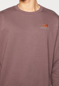 Tröja i dämpad mauve med rund hals och ribbade muddar. Broderad New Balance-logotyp i orange och ljusblått på bröstet.