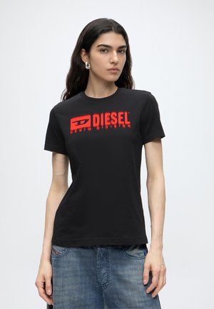 Joven mujer con cabello largo y oscuro, vistiendo una camiseta negra de Diesel con logo rojo y vaqueros azules, sobre un fondo blanco liso.