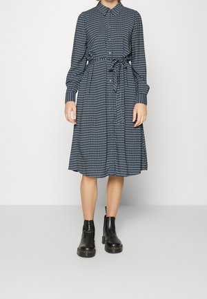 Robe bleu marine avec motif géométrique blanc, manches longues, bouton devant, ceinture à la taille et jupe évasée. Portée avec des bottines noires.