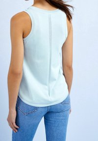 Femme portant un haut sans manches bleu clair avec une couture verticale dans le dos et un jean bleu, vue de dos.