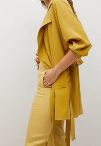 Vêtement d'extérieur jaune moutarde avec un col drapé, des manches larges et une ceinture à nouer à la taille, associé à un pantalon jaune assorti. Texture lisse.