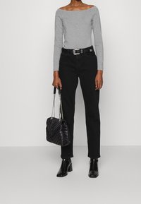 Person som bär en grå off-shoulder topp, svarta högmidjade jeans med bälte, svarta ankelstövlar, och håller en svart kviltad handväska med kedjehandtag.