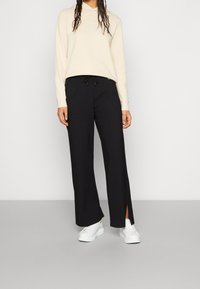 Hoodie beige chiaro con coulisse, logo bianco, abbinato a pantaloni larghi neri con spacchi e indossato con sneakers bianche.