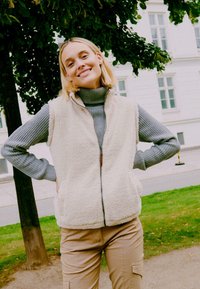 Gilet in pile color crema con zip frontale, superficie texturizzata, abbinato a un dolcevita grigio a coste. Pantaloni cargo color beige completano l'outfit.