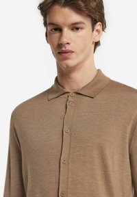 Braunes Strickpoloshirt mit Kragen, ausgestattet mit einer Knopfleiste und langen Ärmeln, aus weichem Material mit glatter Textur.